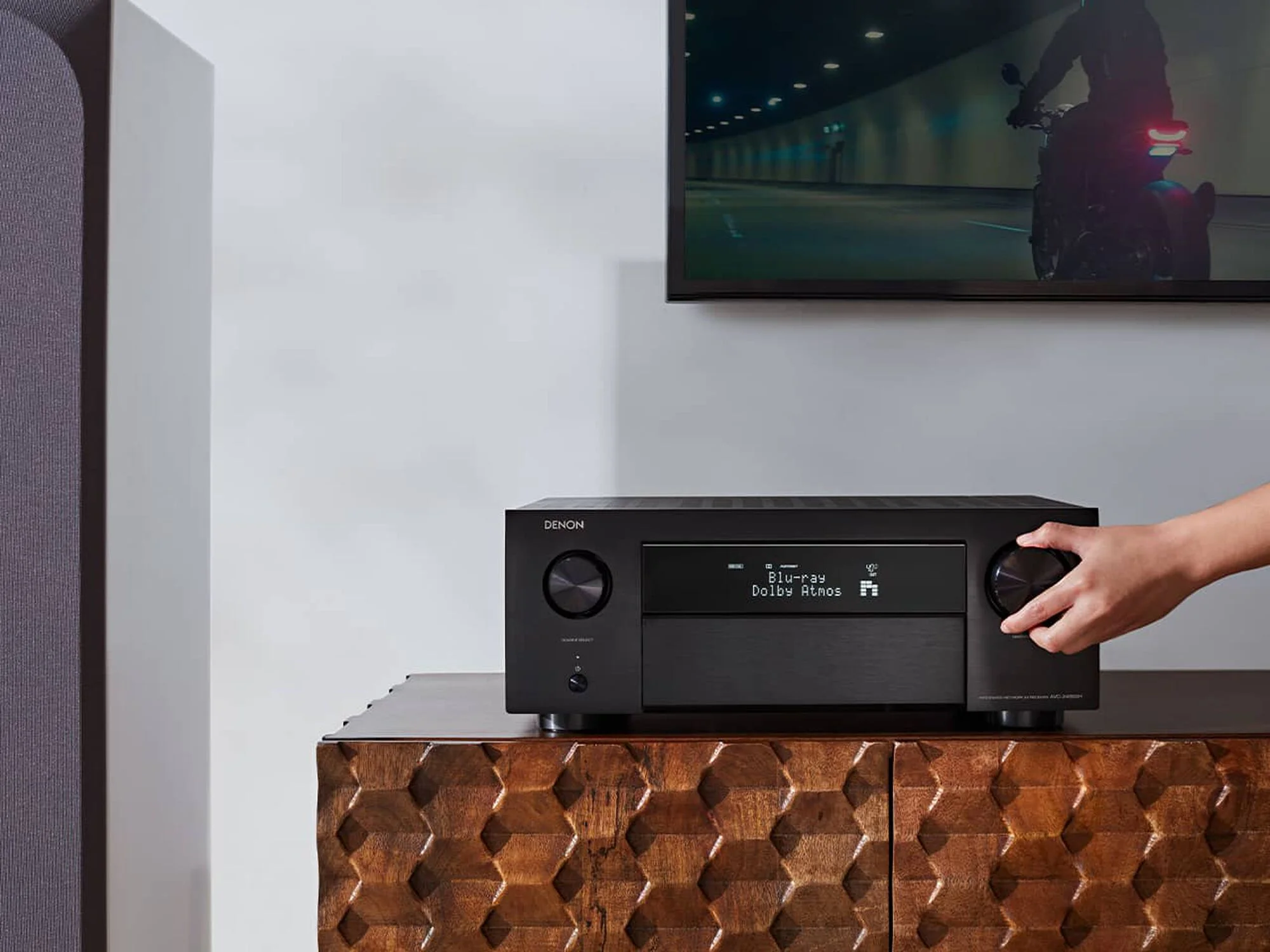 Mano ajustando el control de volumen del Denon AVC-X4800H frente al televisor